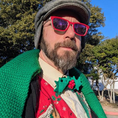 Me, a 46 year old white man with a shaved head, a short cropped brown & grey beard, photographed from the chest up in front of some live oak trees in an office park, wearing a grey flat cap, rectangle glasses with red frames, a green scarf laying on the lapels of an black top coat opened to reveal a holly-shaped bowtie onna cream dress shirt with a very earnestly ugly quilted Christmas music themed vest in red, cream, and green.
