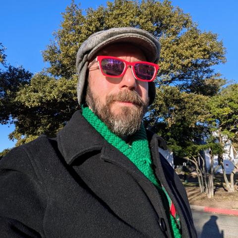 Me, a 46 year old white man with a shaved head, a short cropped brown & grey beard, photographed from the chest up in front of some live oak trees in an office park, wearing a grey flat cap, rectangle glasses with red frames, a green scarf poking out of a black top coat