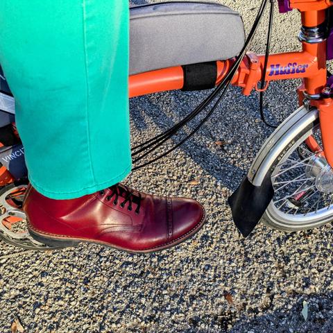 My right foot in a red lace up boot under a green pant leg on the pedal of an orange Brompton folding bike