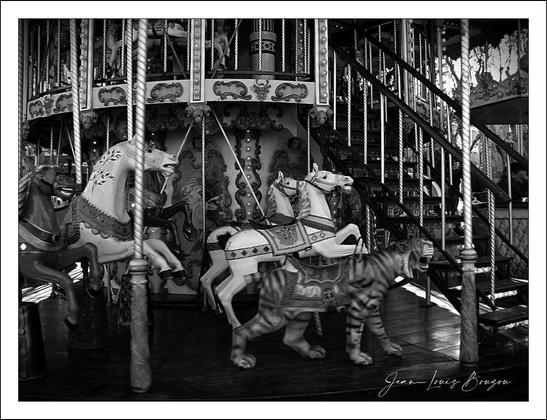 This black and white image depicts a vintage carousel with several intricately designed ride-on figures. There are elaborately decorated horses and a tiger, all positioned on poles. The carousel features ornate detailing, and there's a staircase that leads to an upper level, indicating it's a multi-tiered carousel. The scene is rich in texture and detail, reflecting a nostalgic and classic amusement park atmosphere.