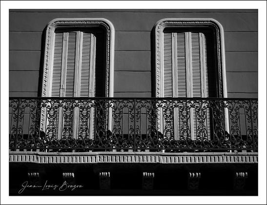
The image depicts a black and white photograph of a building facade featuring two tall, rectangular windows with closed shutters. Each window is framed by ornate moldings. In front of the windows, there is an intricately designed wrought iron balcony railing with decorative patterns. The photo captures the shadows and textures, giving it a classic and architectural feel.