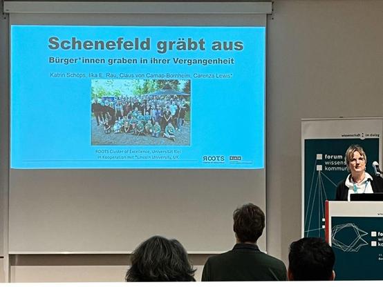 Foto einer Vortragssituation. Groß zu sehen: Eine Leinwand, an die eine PowerPoint-Folie (Titel: "Schenefeld gräbt aus") projiziert wird. Rechts die Referentin am Pult, im Vordergrund die Hintergründe des Auditoriums.