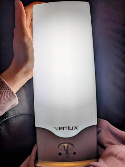 White woman's hand is holding a white rectangular seasonal affective disorder treatment device. It's a very bright lamp. She is holding it on her lap about 12 inches from her face. The photo is taken from her point of view. 