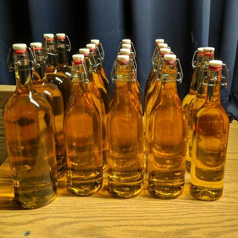 A square cropped photo showing 20.5 liters of wine in 19 bottles as viewed inline with the five rows of bottles. The wine is light amber colored, or probably some wine specific color... On the left there are four 1.5 liter bottles, in the middle are twelve 1 liter bottles, and on the right are two 750ml bottles and one 1 liter bottle. All the bottles have swing tops with white caps and red gaskets, some are cork type wine bottles with a metal band added that allows mounting a swing top.