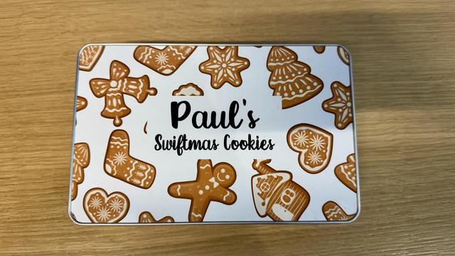 A metal tin featuring the text "Paul's Swiftmas Cookies," decorated with various gingerbread cookie designs, including stars, hearts, and a gingerbread man, set against a white background.