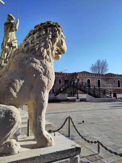 Sculpture of a sitting lion, in white marble, with a curved runic inscription on the shoulder. To the right a mostly empty square, and a wooden bridge in the background.