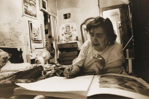 
Foto en blanco y negro de la poeta Alda Merini sentada en una habitación desordenada, escribiendo en un gran libro abierto sobre una cama. La habitación está llena de objetos personales, incluyendo cuadros en las paredes, un ventilador y una cómoda. La mujer parece concentrada en su escritura, y su expresión es seria. La atmósfera de la imagen es íntima y evoca un sentimiento de tranquilidad.