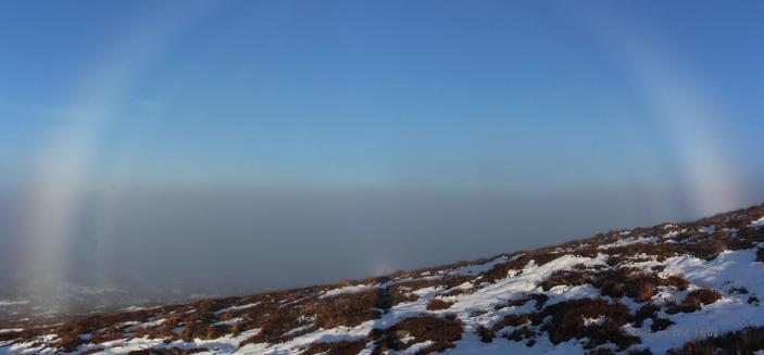 Looking across a sloping hillside with patches of snow.Blue sky, but a layer of cloud below us. There is my shadow on the ground with a glory where is appears on the cloud layer. There is also a double fogbow forming an arc around the glory.