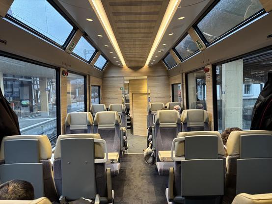 Ein Zugwagen mit Panoramafenstern, gemütlichen Sitzen und hellem Holzinterieur. Die großen Fenster bieten Ausblick auf die winterliche Umgebung.