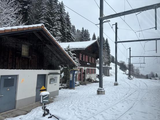 Ein verschneiter, ländlicher Bahnhof mit traditionellen Holzhäusern und roten Fensterläden, umgeben von schneebedeckten Tannen.