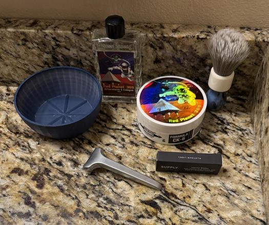 Shaving products on a counter top. Products include a shaving bowl, shaving brush, soap container, aftershave, package of injector razor blades, and an injector type safety razor.