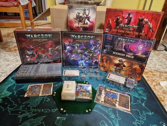 Foto de todo el contenido de mi pledge del juego de mesa Warcrow Aventures. Hay una caja de juego básica, una expansión de historia y verías expansiones de enemigos, héroes, dados y escenografía. Todo ello sobre un tapete verde con un mapa del mundo.
