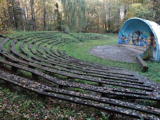 Outdoor amphitheater with wooden seating and a painted stage, surrounded by trees and autumn leaves.
