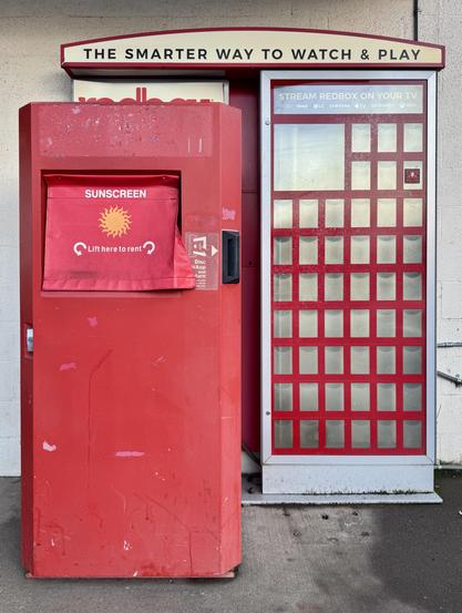 A Redbox vending machine. Screen cover labeled "Sunscreen" is on the left, with a cover that instructs to "Lift here to rent." To the right, there is a grid layout for displaying movies but they are empty.
