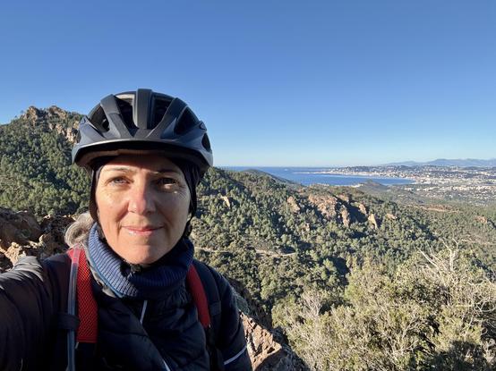 A white woman wearing a helmet and outdoor gear poses at a mountain pass, overlooking a scenic view of green hills and a coastal town under a clear blue sky.