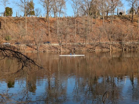 Photo: A sweep-oared pair — two rowers each with a single oar – on a near mirror-still body of water, below a lightly wooded ridge.