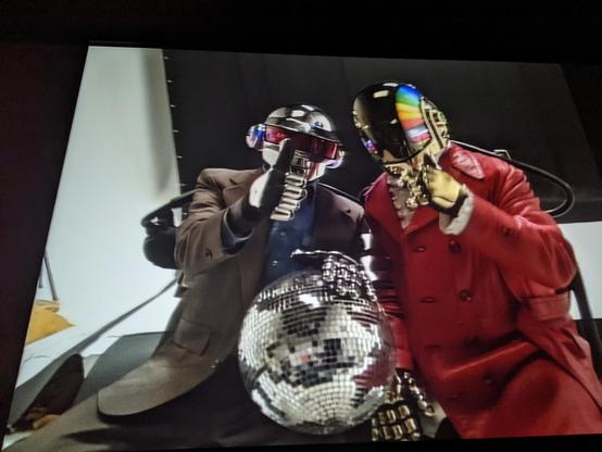 Photo of the end of the film with Daft Punk onscreen in their earlier years. Both are sitting down. Thomas is holding a disco ball wearing a darker suit with the thumbs up sign. Guy-Man is wearing a red jacket & almost forming a thumbs up sign.