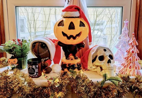 Photo of a decorated shelf. It includes a Sam from Trick 'R' Treat doll wearing a Santa hat and holding a jar candle, a plastic jack 'o lantern wearing a Santa hat, and a plastic skull with a googly eye on it's forehead wearing a Santa hat. Gold tinsel garland and small LED lights hang around the edge.
