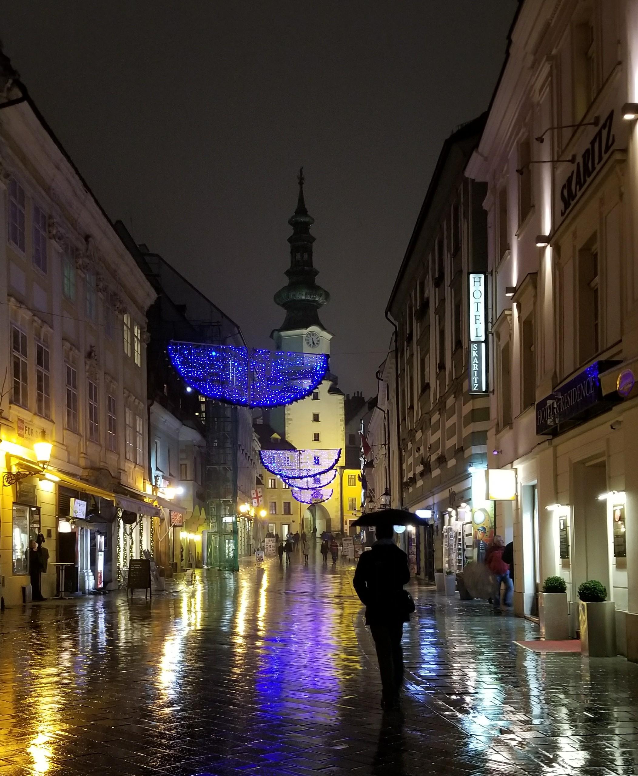 A%20rainy%20night%20of%20old%20town%20Bratislava%20on%20a%20wet%20cobblestone%20pedestrian%20street.%20Either%20side%20of%20the%20street%20has%20older%20buildings%203%20or%204%20stories%20tall%20with%20storefronts%20on%20the%20ground%20floor%20and%20hotels%20or%20apartments%20above.%20Lights%20from%20the%20storefront%20and%20holiday%20lights%20strung%20across%20the%20street%20leave%20%22wet%22%20streaks%20of%20yellow%20and%20purplish%20lights%20on%20the%20ground.%20The%20view%20of%20the%20street%20ends%20with%20a%20pale%20yellow%20clock%20tower%20and%20dark%20spire%20against%20a%20dark%20grey%20sky.