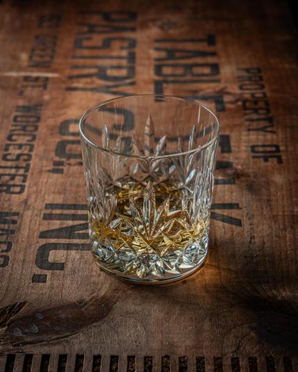 A square photograph of an Edinburgh crystal glass with a finger of amber whisky in it, on the aged wood surface of an old Table Talk Pies promotional case. 
