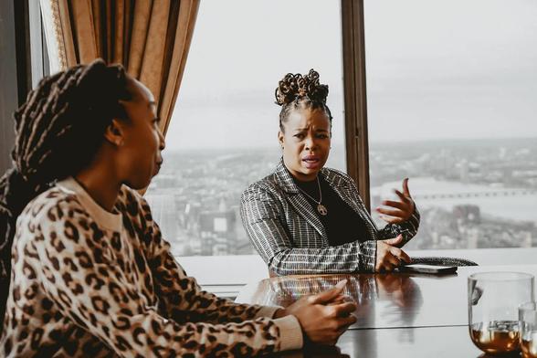 The Ratchet Sage™ and her client, Drudys Ledbetter sit at a table in front of a large window with a city view. Drudys gestures while speaking, wearing a checkered blazer—a solopreneur sharing insights. The Ratchet Sage™ listens attentively in a patterned sweater, as Drudys’ coach. A glass with a drink is visible on the table.