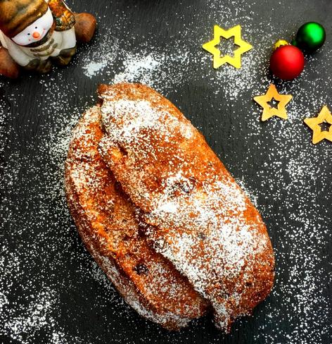 Ein Foodphoto von einem Stollen, der auf einem schwarzen Untergrund liegt. Der Stollen und auch der Untergrund sind mit Puderzucker bedeckt. Links oben im Bild sitzt ein fröhlicher Schneemann und rechts oben liegen 3 goldene Stern, eine rote Weihnachtsbaumkugel und eine grüne Weihnachtsbaumkugel als Dekoration.