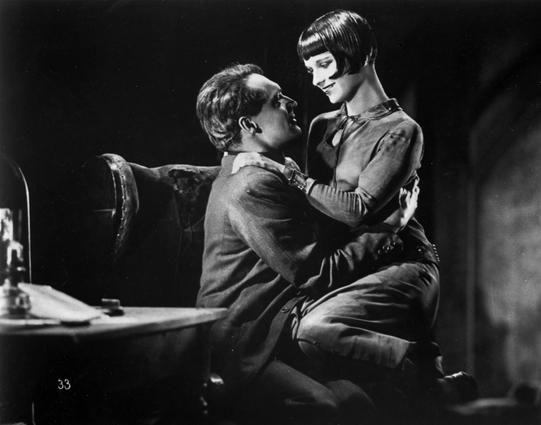 A black-and-white still from the 1929 silent film Pandora's Box features a dramatic and intimate moment between two characters played by Louise Brooks and Francis Lederer. The woman played by Louise Brooks, dressed in a sleek, form-fitting outfit and sporting a short bob haircut, is sitting on the man's lap with her arms gently draped around his shoulders. The man, in a suit, gazes up at her with a look of admiration and longing. The setting appears dimly lit, with shadows and a dark atmosphere contributing to the film noir aesthetic. This visually striking image captures the chemistry and tension between the characters, reflecting the film's themes of passion, desire, and tragedy.