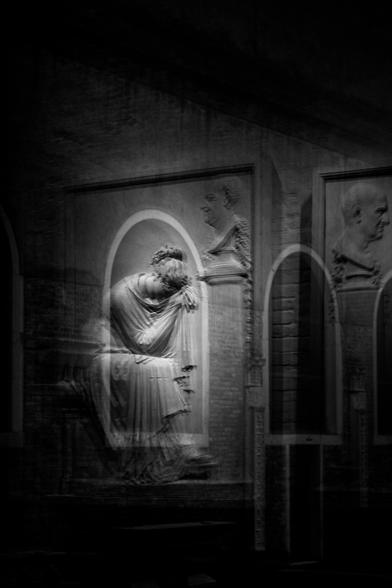 Fotografía en blanco y negro donde se ve el reflejo de una escultura en relieve con posición algo pesarosa y enmarcada por un vano exterior del edificio.