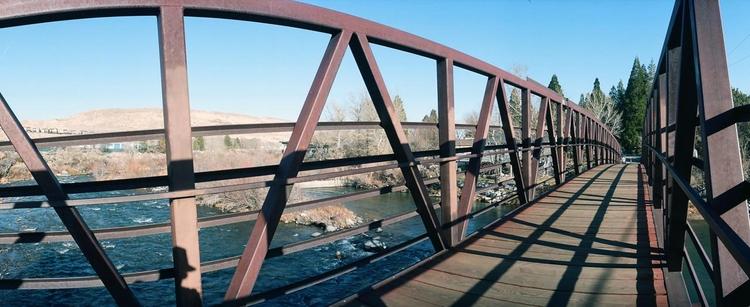 Panoramic photo of a steel and wood footbridge over the Truckee River. The sky is clear blue and the sunlight is strong, so that the iron framework makes a network of sharp shadows.