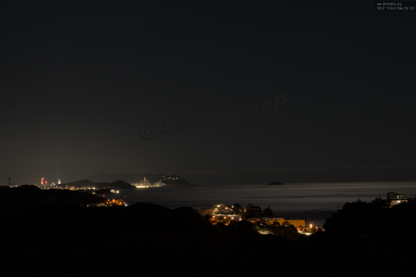 Una impresionante vista nocturna de la costa, donde las luces de Benidorm y otras localidades costeras brillan en el horizonte. La calma del mar, suavizada por la larga exposición, crea un efecto sedoso que contrasta con la intensidad de la iluminación urbana. Una pequeña isla se distingue en la lejanía, añadiendo un punto de interés al paisaje.