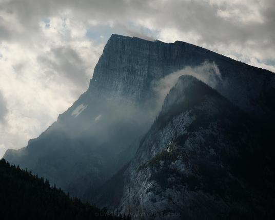An image of crown mountain. The mountain has clouds pressing over its closer ridges as a recent rainfall ends. The blue colour comes from the shadows created by the warm sun backlighting the scene.