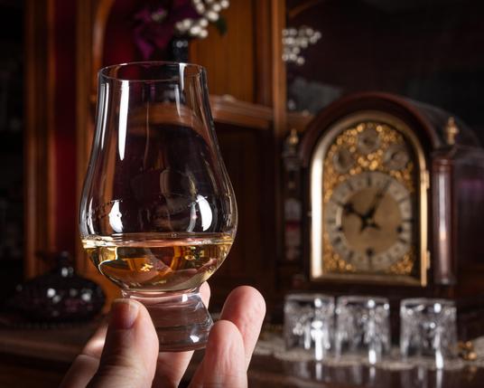 A photograph of the fingertips of a hand holding a Glencairn glass of pale whisky, with an antique clock and some cut crystal glasses on a wooden sideboard behind. The lighting is chiaroscuro and comes from the right-hand side of the scene. 