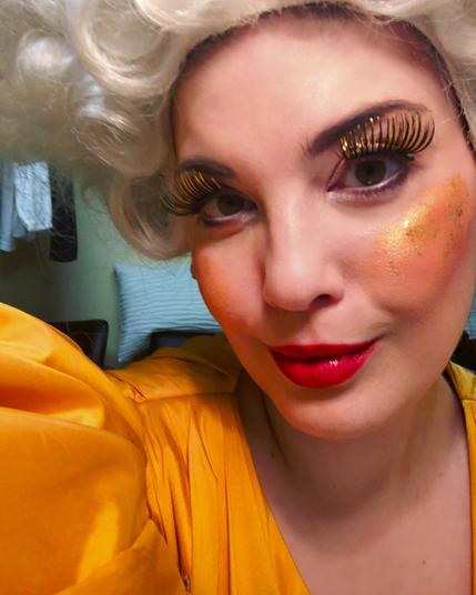 Woman in a yellow ball gown with puffed sleeves is wearing a white wig, red lipstick and enormous gold-tipped eye lashes. She looks into the camera with a smile. Megan Marie Hart backstage in her dressing room between two performances of „Der kleine Prinz“, the German-language version of Pierangelo Valtinoni's opera «Il Piccolo Principe» after Antoine de Saint Exupéry’s «Le Petite Prince». She is in costume, wig and make-up as „Die Eitle“ (La Vanitosa/The Vain Lady). Costume by Camilla Hägebarth, photo by Megan Marie Hart, CC BY-SA4.0.