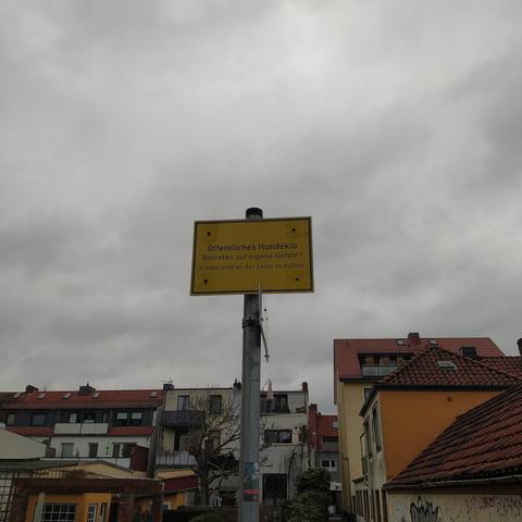 Das obere gelbe Schild aus einer anderen Perspektive vor grauem Himmel und mit Häusern im Hintergrund. 

Beschriftung:
Öffentliches Hundeklo
Betreten auf eigene Gefahr!
Kinder sind an der Leine zu halten. 