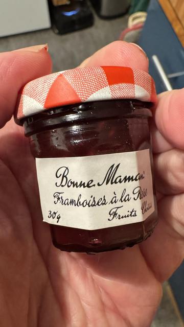 A small pot of red jam held in a hand. It is labelled in cursive French: Framboises à la Rose.