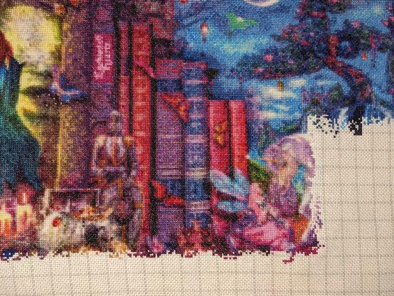 Close up of a very large cross stitch piece. In this section you can see a group of books on a shelf with a treasure chest, armor stand, fairy, and the beginnings of a unicorn in front of them.