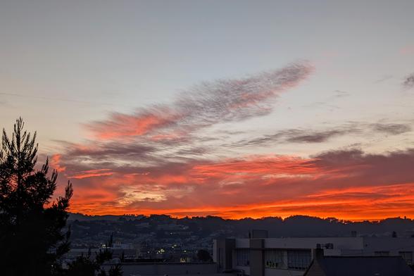Photograph of the sunset over San Francisco Bayview. The lower clouds are lit up intensely orange and salmon, in contrast to their amethyst shadows. Behind the clouds, the sky is a clear pale blue tinted with yellow. CC BY-SA Kate Zimmerman.