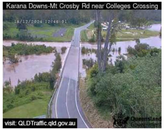 image shows a flooded road in Karana Downs-Mt Crosby Rd near Colleges Crossing in Queensland, Australia. The water level is high, covering the entire road surface and reaching up to the bottom of the guardrails. The water is a murky brown color, suggesting it is carrying sediment and debris. Due to the flooding, the road has been closed by police, requiring motorists to take a lengthy detour. In the background, there are trees and lush green vegetation, indicating a rural area. The image was captured on December 18, 2024, at 12:46:01 PM. 
