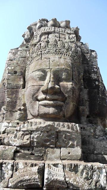 A head on the Bayon at Angkor Thom, possibly depicting Jayavarman VII, the ruler at the time the Bayon was built. It is carved from blocks of grey stone and depicts a broad, smiling face wearing an ornate head dress. The blocks are sized so that the actual face is about two blocks wide and four tall, with the division down the middle of the node. The lower lip is all one block, parenthesised by smaller blocks either side.