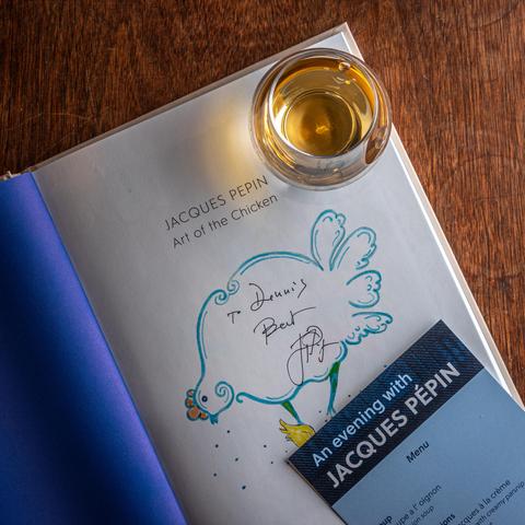 A photograph of a Glencairn glass of whisky sitting on top of an open copy of Jacques Pépin's "The Art of the Chicken", signed "To Dennis, best. Jacques Pépin". Part of a copy of the program/menu for "An Evening with Jacques Pépin" is visible in the lower right corner. 