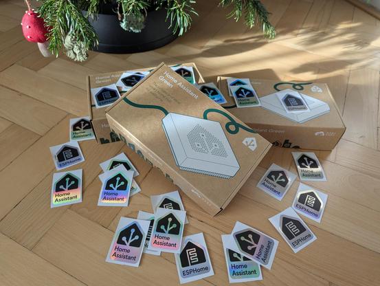 Samolepky ESPHome a Home Assistant, a tři krabice Home Assistant Green na podlaze pod vánočním stromkem. 