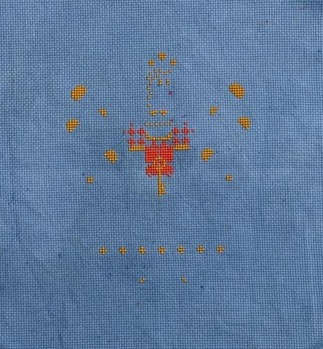 A deep cool blue fabric with yellow cross stitch spots scattered about in a pattern. Red cross stitch areas have started in the center. The final image is still unclear.