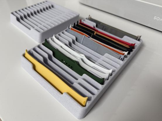 A neat, organized display of colorful watch bands stored in a gray tray with multiple compartments. The bands include silicone, fabric, and nylon materials in various colors like yellow, green, white, red, and gray, showcasing a stylish collection for interchangeable smartwatches.