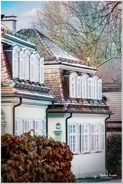 Das Foto zeigt ein elegantes, historisches Haus mit Schindeldach, teilweise mit Schnee bedeckt. Es verfügt über schöne weiße Fensterläden und Erkerfenster. Im Vordergrund befinden sich braune Pflanzen, im Hintergrund kahle Bäume und ein weiteres Gebäude.
