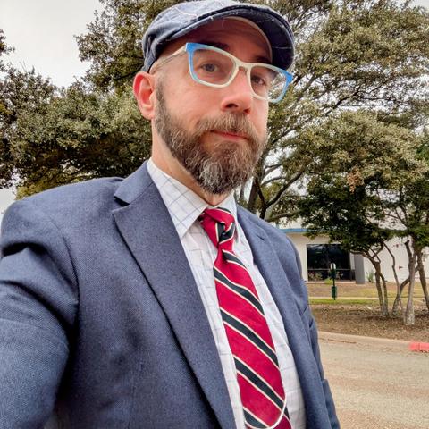 Me, a 46 year old white man with a shaved head, a short cropped brown & grey beard, 

photographed from the chest up in front of some live oak trees in an office park,

wearing

a rectangle glasses with white and blue frames, a white dress shirt wuth a blue windowpane pattern, a deep red necktie with navy stripes in a trinity knot under a navy blue blazer with a medium blue flat cap