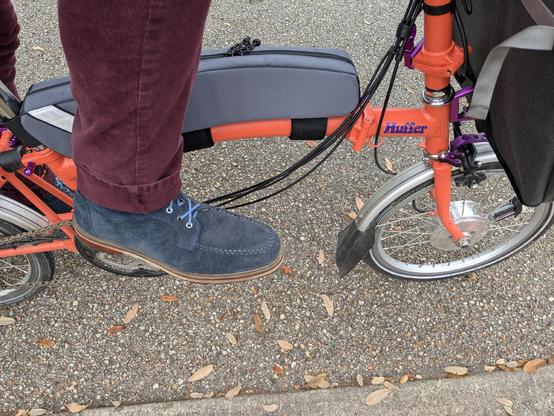 My right foot in a blue suede moccasin toe boot with light blue laces under a dark red corduroy pant leg on the pedal of an orange Brompton folding bike with Huffer, its name, in purple text on the main tube 