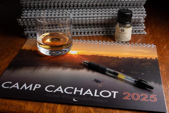 A photograph of a doubles glass of whisky sitting on top of an Camp Cachalot 2025 photo calendar, in front of a stack of calendars. A pen sits on top of the calendar as well, ready to sign it, and the small bottle the whisky came in, labeled Glen Scotia, is near the stack on the right. 
