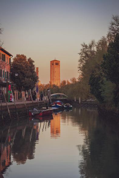 Imagen vertical donde se ve un canal con algunas barcas y la torre de la basílica Santa María Asunta con la luz del atardecer.