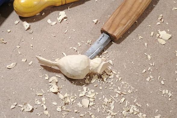 The rough shape of a small bird with beak wide open sits among a pile of wood chips with a carving knife holding it up a bit.