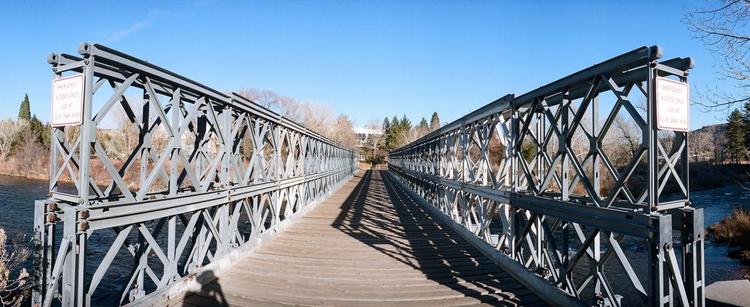 Panoramic photo with a view straight down the middle of an iron and wood footbridge. It is very symmetrical. Signs on either side warn that the bridge is for “Emergency access only. Use at your own risk”.

